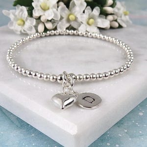 May include: A silver beaded stretch bracelet featuring two dangling charms: a smooth, polished silver heart and a small, round silver disc engraved with the capital letter D. The bracelet is constructed from small, uniform metallic spheres.