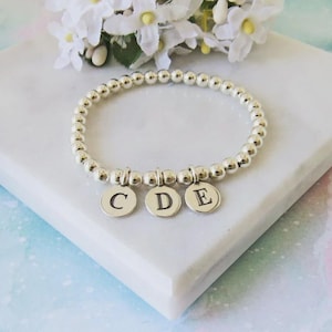 Pode incluir: Pulseira de contas prateadas com três pingentes redondos gravados com as letras C, D e E. A pulseira é exibida em uma superfície de mármore branco com flores brancas ao fundo.