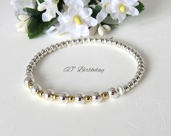 Könnte beinhalten: Ein elastisches Armband aus kleinen metallisch silbernen Perlen, die mit größeren silber- und goldfarbenen Perlen durchsetzt sind. Der Text 60th Birthday ist mittig in der kreisförmigen Anordnung der Perlen platziert, die ein Muster bilden.