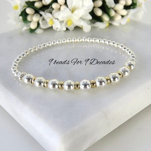 May include: A stretch bracelet featuring small silver-colored beads and larger silver and gold-colored beads arranged in a circular pattern. The text 9 beads For 9 Decades is printed in black script in the center of the bracelet design.