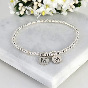 May include: A silver beaded bracelet with two round charms, each displaying the letter "M". The bracelet is displayed on a white marble surface, with white and yellow flowers in the background. The bracelet is made of small, round beads.