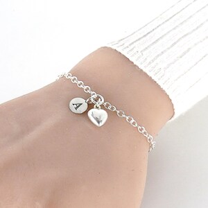 May include: A silver-toned chain bracelet featuring two dangling charms: a small circular disc engraved with the capital letter A and a smooth, polished heart-shaped charm. The bracelet is constructed with a simple link chain design.