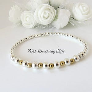May include: A stretch bracelet composed of small silver-toned beads with alternating gold-toned accents. The text 70th Birthday Gift is centred above the bracelet. The design features a repeating pattern of metallic spheres arranged in a circular loop.