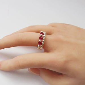 May include: A silver ring with two bands, each decorated with small silver beads. The top band has a row of bright red beads. The ring is worn on a finger, highlighting its design and colour contrast.