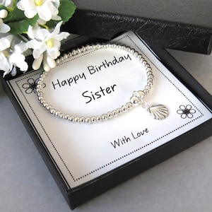 May include: A silver beaded bracelet with a small seashell charm, presented in a black gift box on a white card featuring the text Happy Birthday Sister With Love, decorated with black line-art flowers and accompanied by white flowers.