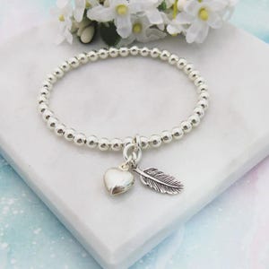 May include: Silver beaded bracelet with a heart and feather charm. The bracelet has small, round beads and a dangling heart-shaped charm and a feather charm. The bracelet is displayed on a white marble surface with white flowers in the background.