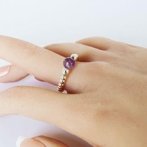 May include: A stretch ring featuring a single round purple amethyst bead centred among multiple small, polished silver-toned metallic beads. The ring is constructed with a flexible band design that allows it to fit comfortably around a finger.