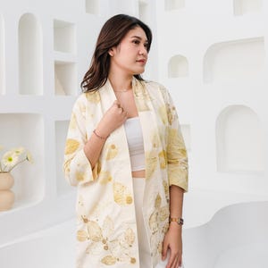 May include: Cream-colored kimono-style jacket with a botanical leaf pattern in shades of gold and brown. The jacket is open-fronted and has three-quarter sleeves. The model is wearing a white top and pants.