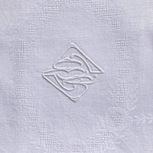 May include: A white, textured fabric with a repeating geometric pattern. The center features an embossed, abstract design within a diamond shape. The overall effect is a subtle, elegant texture, suitable for linens or decorative textiles.
