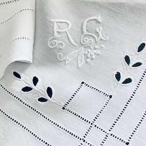 May include: White linen napkin with embroidered initials "RG" and floral accents. Features cutwork leaf designs and geometric patterns. The napkin is folded, showcasing a detailed, elegant design.