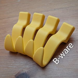 Könnte beinhalten: Vier gelbe Kunststoffhaken mit wellenförmigem Design. Die Haken sind in einer Reihe angeordnet und zum Aufhängen von Gegenständen bestimmt. Der Text "B-ware" ist unten rechts zu sehen. Jeder Haken ist etwa 10 cm lang.