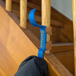 Peut inclure: Un crochet en plastique bleu avec une partie supérieure incurvée et une section verticale plate portant le nom Nikolaus en écriture jaune. Le crochet est fixé à un balustre d'escalier en bois avec deux sangles en daim beige.