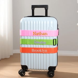 May include: A light blue rolling suitcase with three colorful straps. The straps are pink, green, and orange, each embroidered with a name: Nathan, Sebastian, and Brooklyn. The suitcase has a retractable handle and four black wheels.
