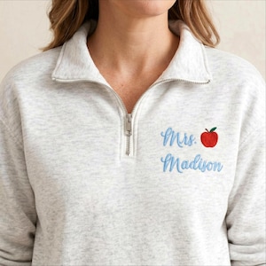 May include: A light gray sweatshirt with a quarter-zip neckline. The sweatshirt features the embroidered text "Mrs. Madison" in light blue, with a red apple graphic. The zipper is silver.