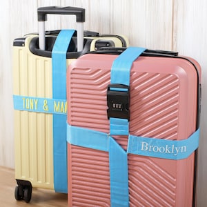 May include: Two suitcases, one cream and one pink, secured with light blue luggage straps. The cream suitcase has the text "TONY & MA" and the pink suitcase has "Brooklyn" on the straps. Both have black combination locks.