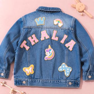 May include: A blue denim jacket featuring the name THALIA in large pink letters across the back. The jacket is decorated with embroidered patches including a blue crown, a rainbow, a unicorn head, a yellow butterfly, and a blue polka dot bow.