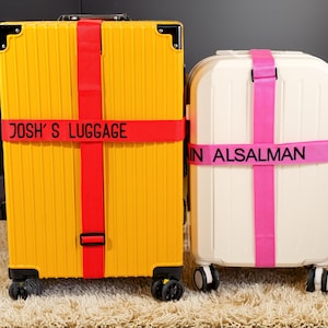 Puede incluir: Dos maletas, una amarilla y otra blanca, están aseguradas con correas. La maleta amarilla tiene una correa roja con "JOSH'S LUGGAGE" impreso. La maleta blanca tiene una correa rosa con "IN ALSAlMAN" impreso. Ambas están sobre una alfombra beige.