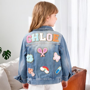 Pode incluir: Casaco de ganga azul claro com aplicações bordadas coloridas. As costas do casaco apresentam o nome "CHLOE" em letras grandes e multicoloridas. Outras aplicações incluem uma borboleta, raquetes de ténis, um cogumelo, uma nuvem, um arco-íris e uma cápsula.