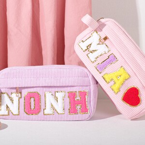 Peut inclure: Deux trousses de maquillage en velours côtelé rose clair. L'une est rectangulaire avec le nom "NONH" en lettres blanches, roses et dorées. L'autre est en forme de trousse avec le nom "MIA" en lettres blanches, violettes et jaunes, et un cœur rouge.