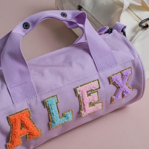 Puede incluir: Una bolsa de deporte lavanda con el nombre "ALEX" en letras bordadas de colores. La bolsa tiene una asa a juego y una segunda bolsa blanquecina es visible en el fondo. Las bolsas están sobre una superficie rosa.