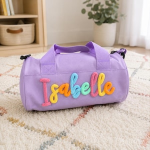 May include: A lavender cylindrical duffel bag featuring the name Isabelle in raised, multi-colored yarn lettering. The bag includes two top handles, a main zippered compartment, and black hardware attachments for a shoulder strap on each side.