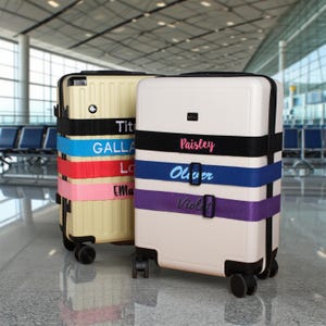 Può includere: Due valigie con ruote con cinturini per bagagli personalizzati. Una valigia è beige, l'altra è bianca. I cinturini sono neri, blu, rossi e rosa su una valigia e neri, blu e viola sull'altra. I nomi sono stampati sui cinturini.