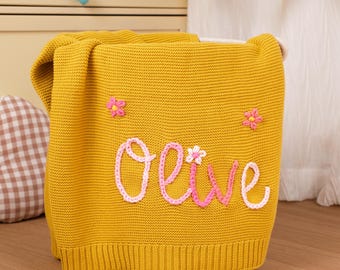 May include: Mustard yellow knitted blanket with the name "Olive" embroidered in pink and white. The blanket features two embroidered pink flowers. The blanket is folded over a white storage container.