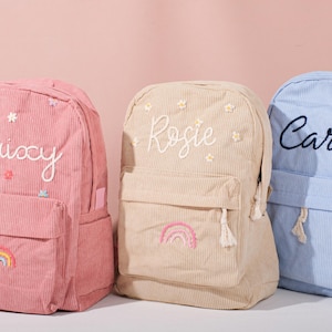 Könnte beinhalten: Drei Cordrucksäcke in Rosa, Beige und Blau. Jeder Rucksack hat eine Vordertasche und ist mit einem Namen in Schreibschrift bestickt: Laixy, Rosie und Cara. Die rosa und beigen Rucksäcke haben ein Regenbogen-Design.