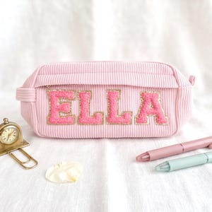 Peut inclure: Une trousse à crayons rose clair côtelée avec le nom ELLA en grandes lettres roses texturées à bordure dorée scintillante. La trousse est accompagnée d'une petite horloge dorée, d'un stylo rose et d'un stylo vert clair.