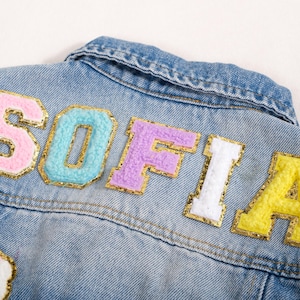 Könnte beinhalten: Nahaufnahme einer hellblauen Jeansjacke mit dem Namen "SOFIA" in farbenfrohen, strukturierten Aufnähern. Die Aufnäher sind goldfarben umrandet und weisen Pastellfarben wie Rosa, Blau, Lila, Weiß und Gelb auf. Ein Regenbogen-Aufnäher ist unten sichtbar.