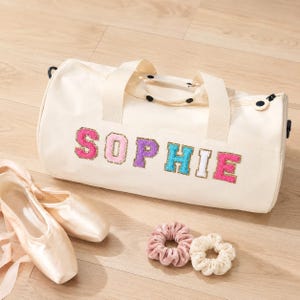 Può includere: Una borsa sportiva in tela color crema con il nome SOPHIE in lettere colorate e testurizzate in stile varsity. La borsa include due manici superiori e una chiusura a zip. Accanto si trovano un paio di scarpette da danza e due elastici.
