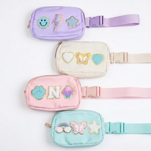 Puede incluir: Cuatro riñoneras coloridas en tonos pastel de lavanda, crema, rosa y turquesa. Cada riñonera presenta parches decorativos, incluyendo una carita sonriente, un rayo, una estrella, un corazón, una mariposa y un arcoíris. Se ven correas ajustables y cierres de cremallera.