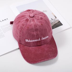 Puede incluir: Gorra de béisbol granate con visera curva. La gorra presenta el texto "Mohammed Jassim" bordado en blanco. Está hecha de un material suave y texturizado y tiene un botón en la parte superior.