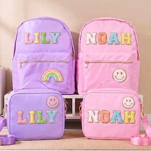 Könnte beinhalten: Zwei Rucksäcke und zwei Lunchpakete in aufeinander abgestimmten Farben. Ein Set ist lavendelfarben mit dem Namen "LILY" und einem Regenbogen-Patch. Das andere Set ist rosa mit dem Namen "NOAH" und einem Smiley-Patch. Alle Taschen haben goldene Reißverschlüsse.