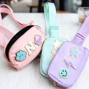 Op de afbeelding: Drie kleurrijke heuptasjes in roze, mintgroen en lavendel. Elke tas heeft een ritssluiting, een verstelbare riem en decoratieve patches, waaronder letters, een pootafdruk, sterren, een bliksemschicht, een cupcake en een smiley.