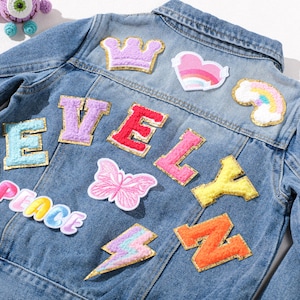 May include: A light blue denim jacket decorated with vibrant patches. Patches include a crown, heart, rainbow, butterfly, lightning bolt, and letters spelling "EVELY" and "PEACE". A fashionable and customisable piece of clothing.