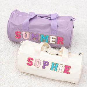 May include: A lavender duffel bag with the word SUMMER in colorful glittery varsity-style letters and a white duffel bag with the word SOPHIE in colorful glittery varsity-style letters, both featuring top handles and a zippered main compartment.