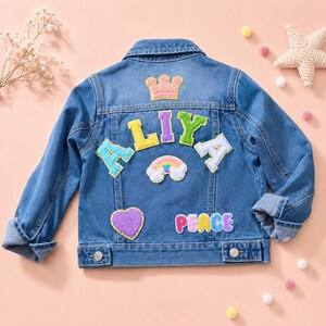 May include: A blue denim jacket featuring colorful embroidered patches on the back, including a pink crown, the name ALIYA in multi-colored letters, a rainbow, a purple heart, and the word PEACE in pastel-colored letters with a white outline.