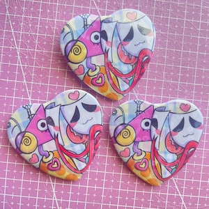 May include: Three heart-shaped badges with cartoon characters. Each badge features a light blue background with a cartoon face, a pink snail, and red and orange tentacles. The badges are on a pink grid surface.