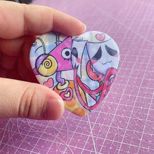 May include: Heart-shaped button with a colorful cartoon design. The button features a pink, yellow, and blue color scheme with whimsical characters and patterns. The button is held in a hand.