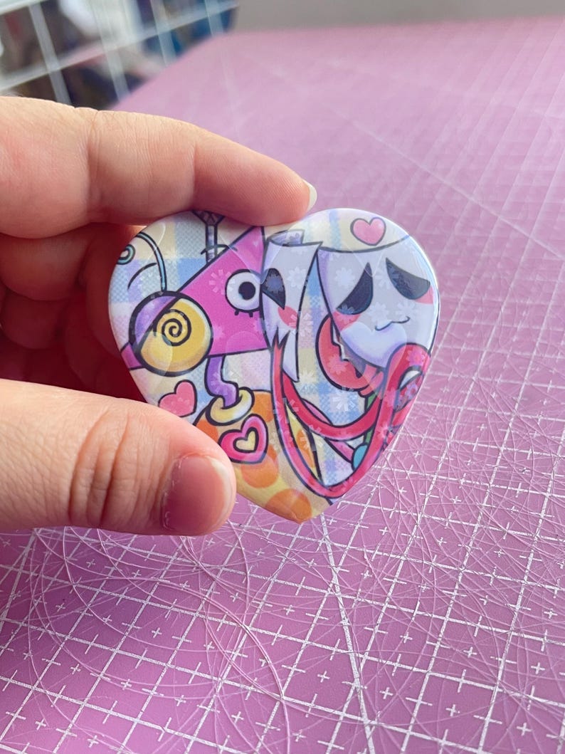 May include: Heart-shaped button with a colorful cartoon design. The button features a pink, yellow, and blue color scheme with whimsical characters and patterns. The button is held in a hand.