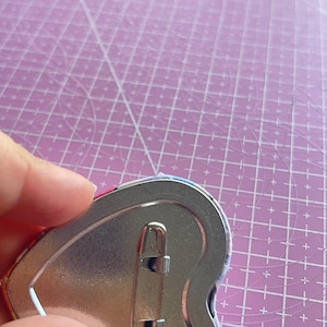 May include: A heart-shaped pinback button with a silver metal backing and a safety pin clasp. The button's edge shows a glimpse of a colorful design. The button is held against a pink cutting mat with a grid pattern.