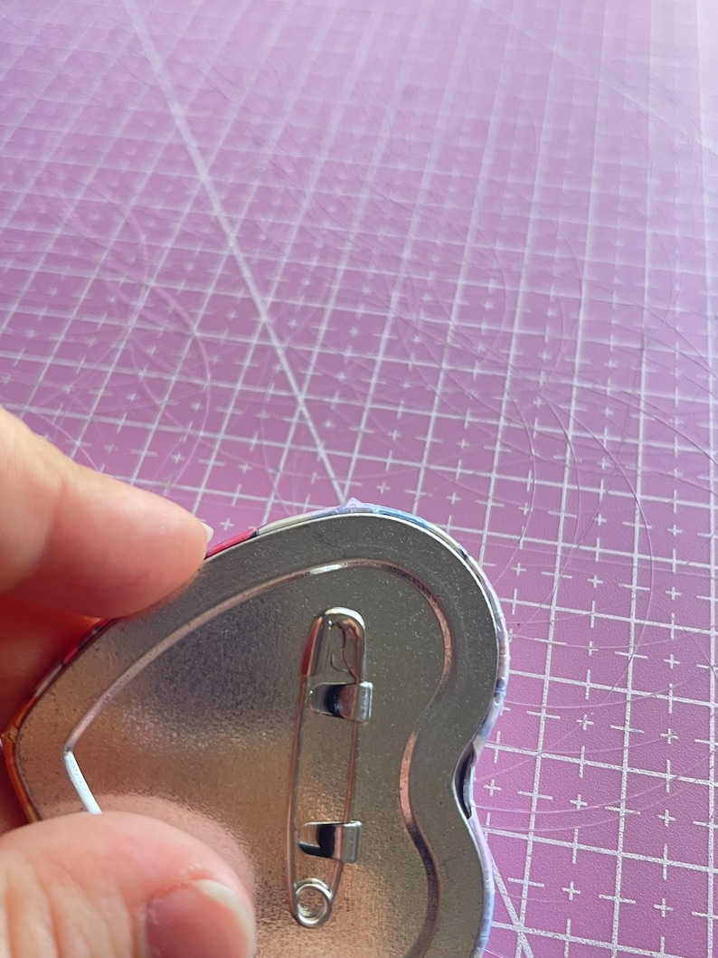 May include: A heart-shaped pinback button with a silver metal backing and a safety pin clasp. The button's edge shows a glimpse of a colorful design. The button is held against a pink cutting mat with a grid pattern.