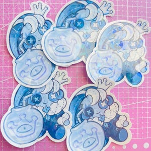 May include: A collection of blue stickers featuring a cartoon character with a crown and a fishbowl. The stickers have a holographic effect and are placed on a pink cutting mat. The character has a button eye and a cross-stitch design.