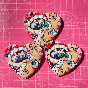 May include: Three heart-shaped pins with a cartoon face design. The faces have large blue and green eyes, a red and white mouth, and a tan skin tone. The pins are on a pink cutting mat.