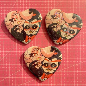 May include: Three heart-shaped buttons with cartoon character illustrations. The buttons feature a pink, black, and red color scheme. The background is a pink grid.