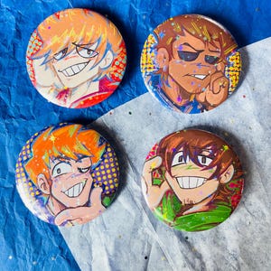 Puede incluir: Cuatro chapas redondas con alfiler que presentan ilustraciones de personajes de estilo anime con superposiciones de purpurina holográfica en forma de estrella. Cada chapa muestra una expresión facial única y una paleta de colores vibrantes en tonos naranja, marrón y verde.