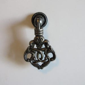 May include: A silver-coloured metal drawer pull with an elaborate, scrollwork design. The pull has a circular backplate and a decorative handle with a detailed, symmetrical pattern. The hardware is likely for a cabinet or drawer.