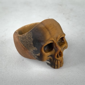May include: A wooden skull ring, intricately carved with a detailed skull design. The ring is predominantly brown with darker brown and black shading. The eye sockets and nose are clearly defined.