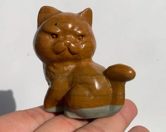 May include: A brown cat figurine with a grumpy expression. The cat is carved from a polished material, with a tail curled to the side. The base of the cat is a light gray color. A decorative item.